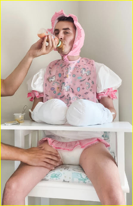 Adult baby DiaperKing in tragic pink bib and bonnet being bottle-fed by daddy